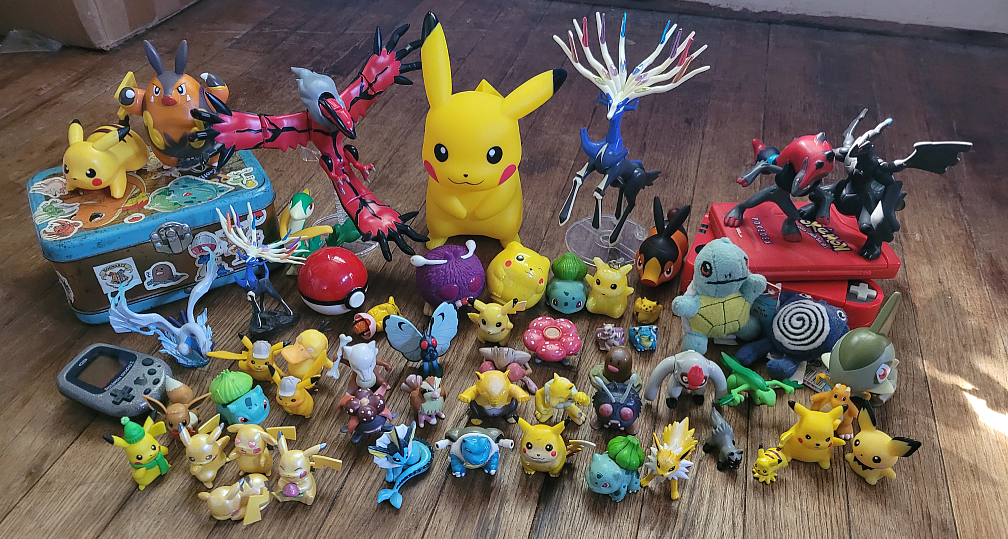 Assorted Pokemon toys, mostly small plastic figurines. There's also a couple Pokedexes, a Pikachu 2 GS, the Soul Silver Lugia figurine, bigger posable Xerneas and Yveltal figurines, a bigger Pikachu, and a metal box featuring Charmander and covered in Pokemon stickers.